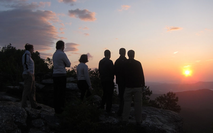 A group of people stand on an overlook and watch the sunset. They appear to be at high elevation in a mountainous landscape, and the sky is lit in soft hues of blue, yellow, orange, pink and purple.